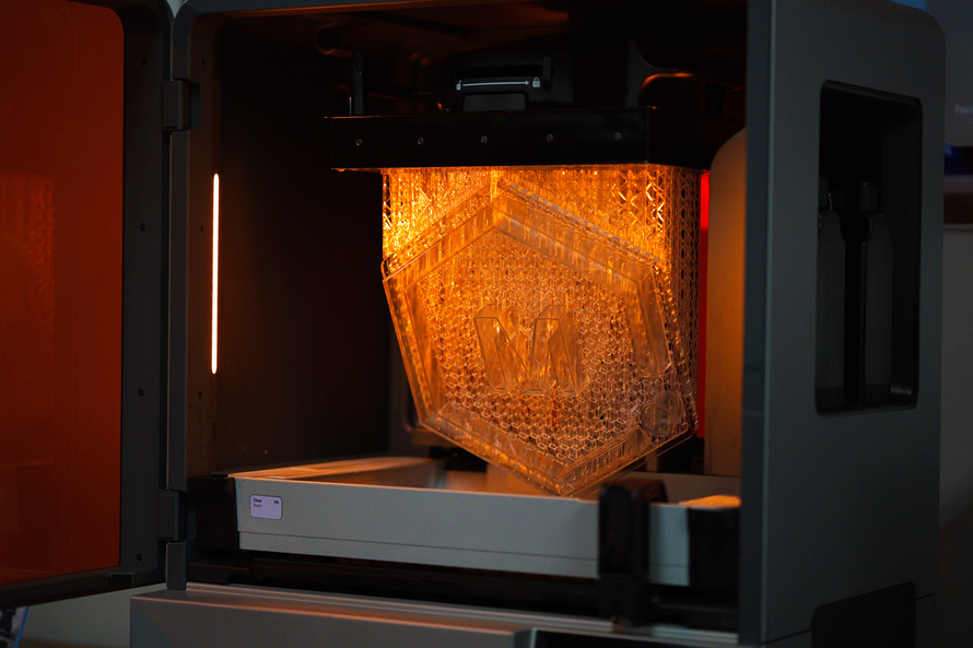 A Clear MH Logo lit by Orange Light Suspended Above a Vat of Clear Resin inside a Formlabs Form 4L
